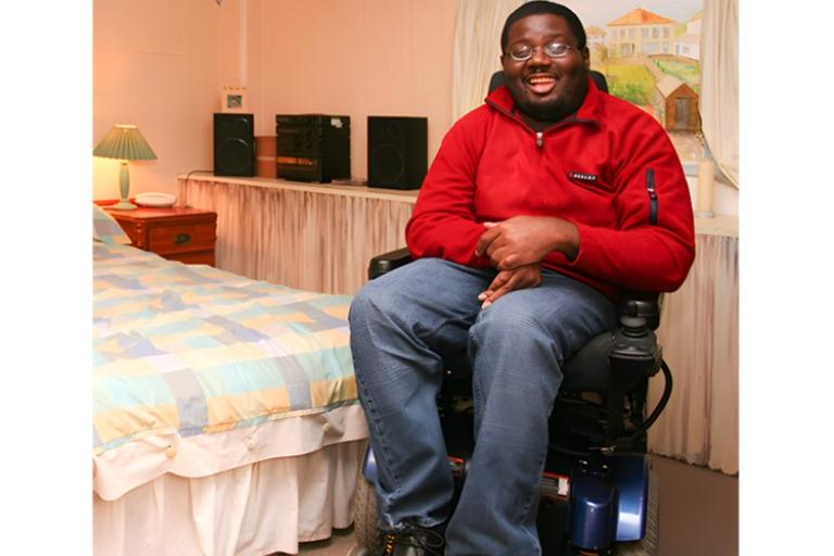 A man in a red jacket sitting in a motorized wheelchair in a cozy bedroom.