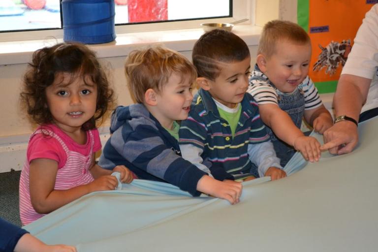 Four nursery aged children holding a stretchy fabric and smiling.