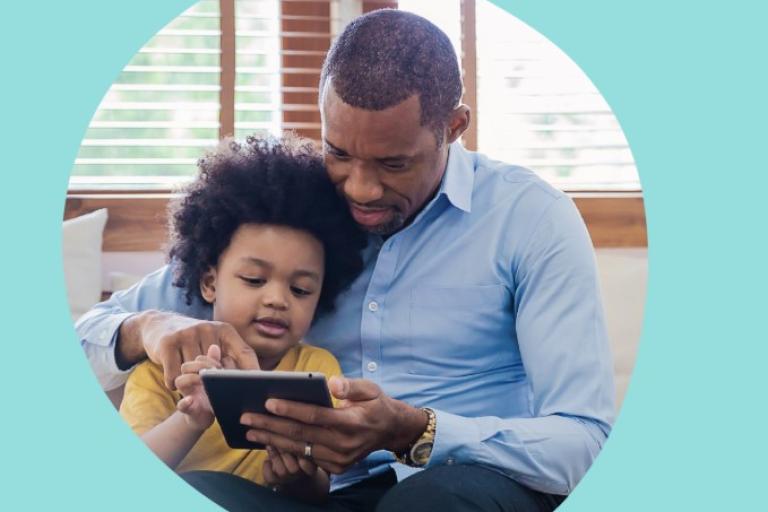 A man and child sitting close together in a bright room looking at a tablet