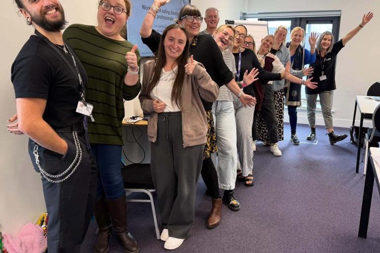 A line of adults standing in a classroom setting smiling and raising their arms for the photo.