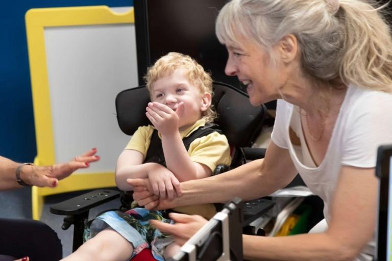 A smiling child in a modified chair holding the arm of a smiling woman.