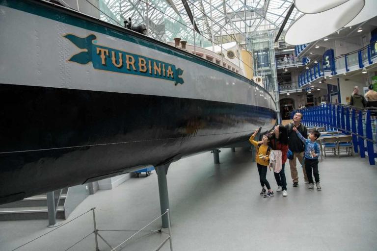 A family standing next to and look at a large shiny long boat with the word Turbinia written on it. The boat is inside a large bright room with a glass ceiling at the Discovery Museum. 