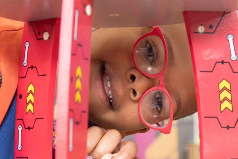 A child wearing glasses looking through a wooden toy space rocket