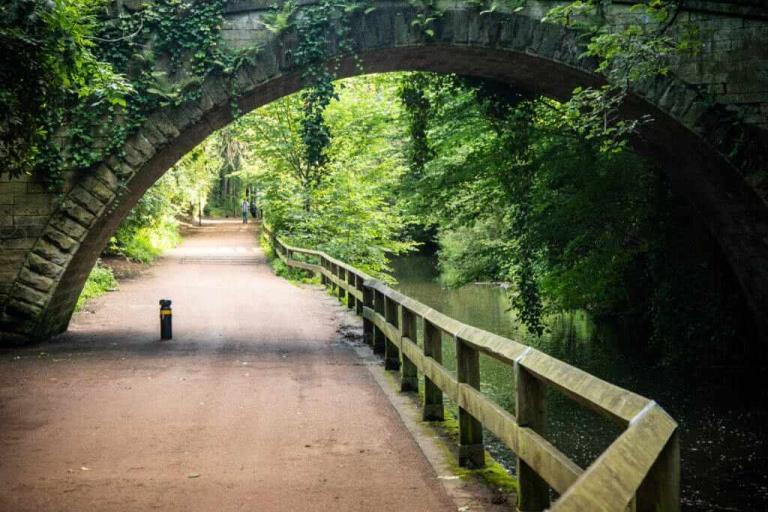 Photo of a wide path alongside a river going under a stony bridge.