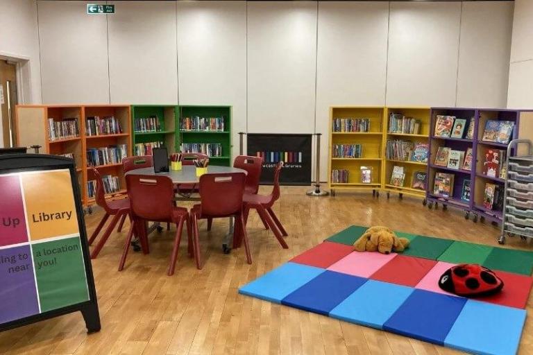 A pop up library in a large room. There are short colourful shelves filled with books, table and chairs, and a simple play area for children