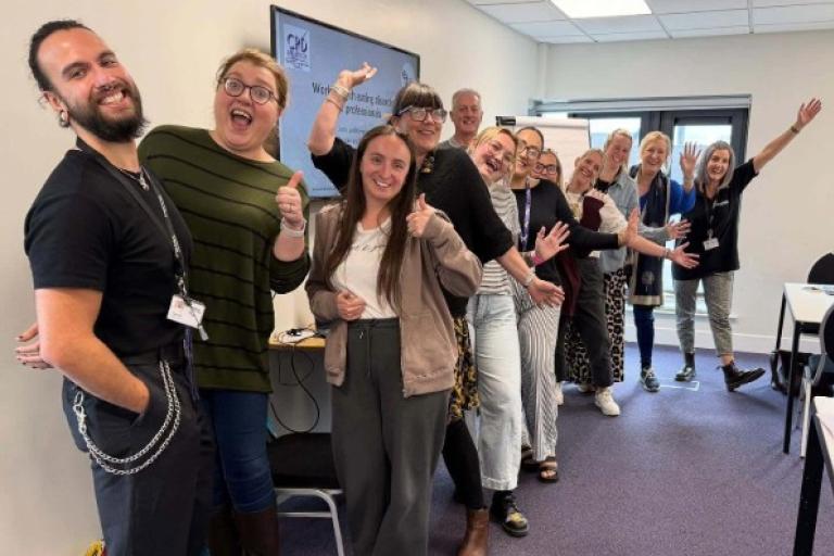 A line of adults standing in a classroom setting smiling and raising their arms for the photo.