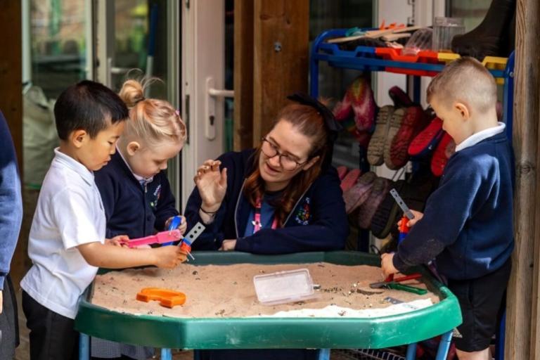 Three young children and an adult playing with a tray filled with sand.