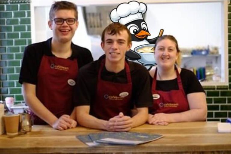 3 young people smiling and wearing aprons with a cartoon penguin wearing a chef's hat in the background