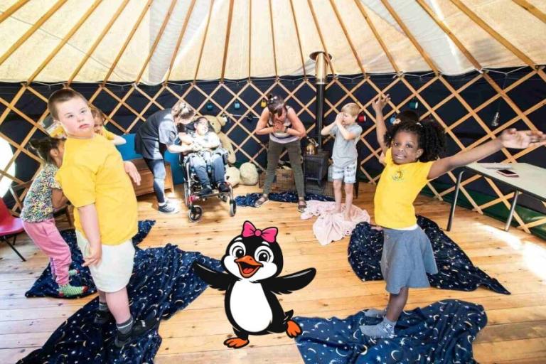 Young children standing on blankets in a round space. A cartoon penguin with a pink bow stands in the middle.