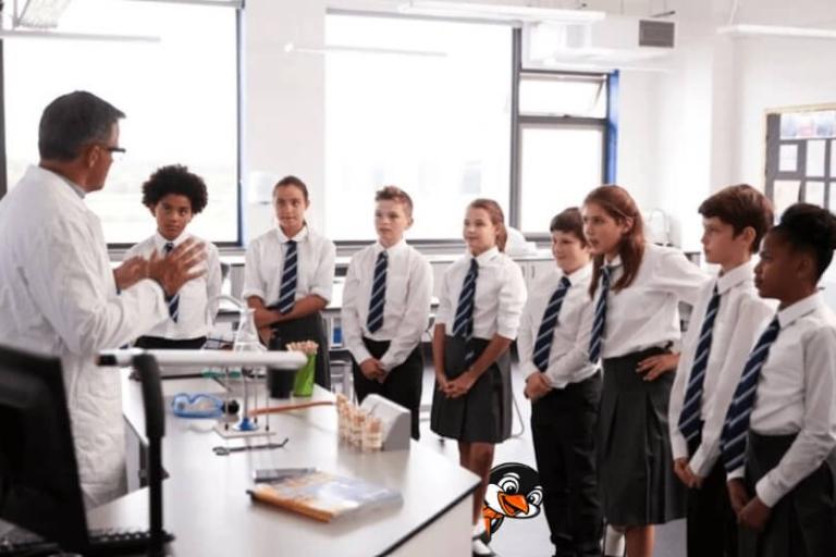A group of secondary school students standing in front of their teacher. A cartoon penguin wearing an orange tie is hiding behind one of the student's legs. 