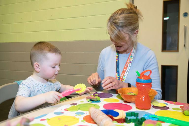 Family practitioner and a young child sat at a table with colourful table cloth playing with playdoh and playdoh tools