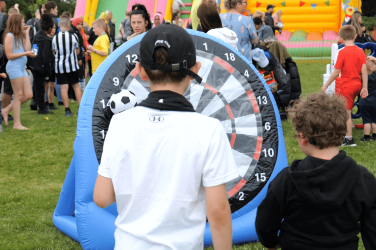 Family fun day outdoors with bouncy castles, lawn games and lots of families in the background and two boys playing with an inflatable football target in the foreground